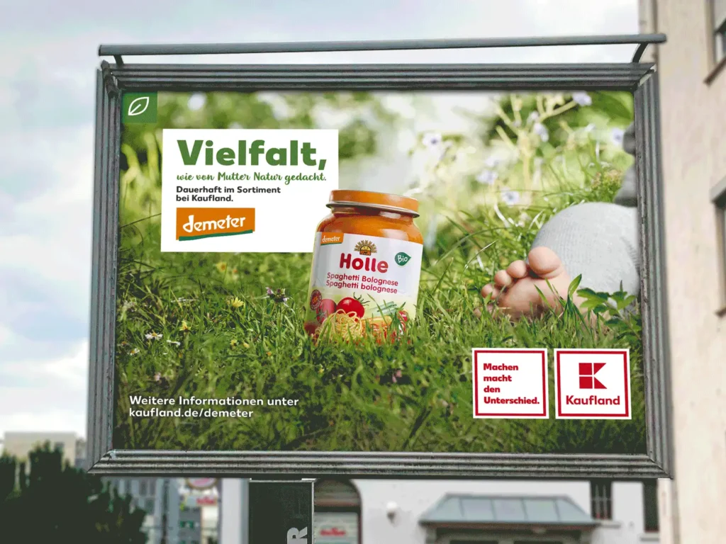  Ein Großen OOH Plakat mit einem hochwertigen Holle Demeter Gläschen auf einer sehr Artenreichen gesunden grünen Wiese. Ein sitzendes Baby sieht man rechts im Anschnitt. Auf dem Plakat steht in einem weißen Kasten der ein wenig von dem nach höchsten Standarts produzierten Produzieren Produkt verdeckt wird: Vielfalt, wie von Mutter Natur gedacht. Unten rechts ist das Kaufland Logo zu sehen 