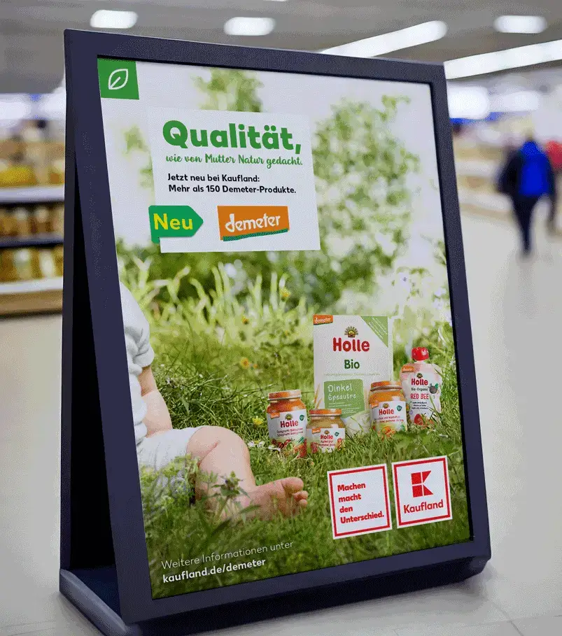 POS Aufsteller bei Kaufland zeigt die Hochwertigen Packshots der Holle Demeter Babynahrung auf einer sehr Artenreichen grünen Wiese. Ein sitzenden Kleinkind sieht mann man im Anschnitt so dass der Fokus auf der Produkten liegt