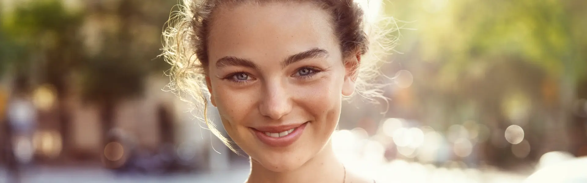 Header Junge lächelnde Frau in einem Frühlingshaften Urbanen Umfeld das Unscharf im Hintergrund zu erkennen ist