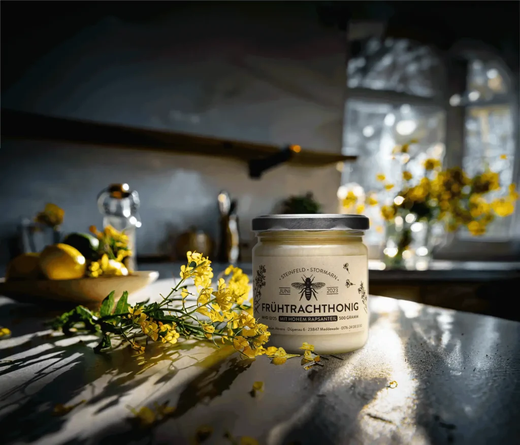 Recyclingpapier für Verpackungen Designlösungen für regionale Honigprodukte. Das Pfandglas steht in einer Lichtdurchfluteten Küche. Um die Hochwertigkeit des Produkts und dessen Inhaltsstoffe zu unterstreichen liegen gesunde Raps Ähren aus Biologischem Anbau neben dem Glas