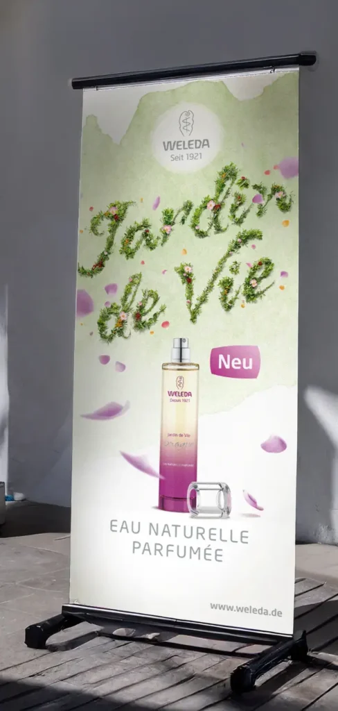 Markenentwicklung für nachhaltige Produkte Messe Roll Up mit einem aus Blumen gelegten Schriftzug, der Jardin de Vie zeigt. Ein paar Blätter fliegen leicht durch die Luft